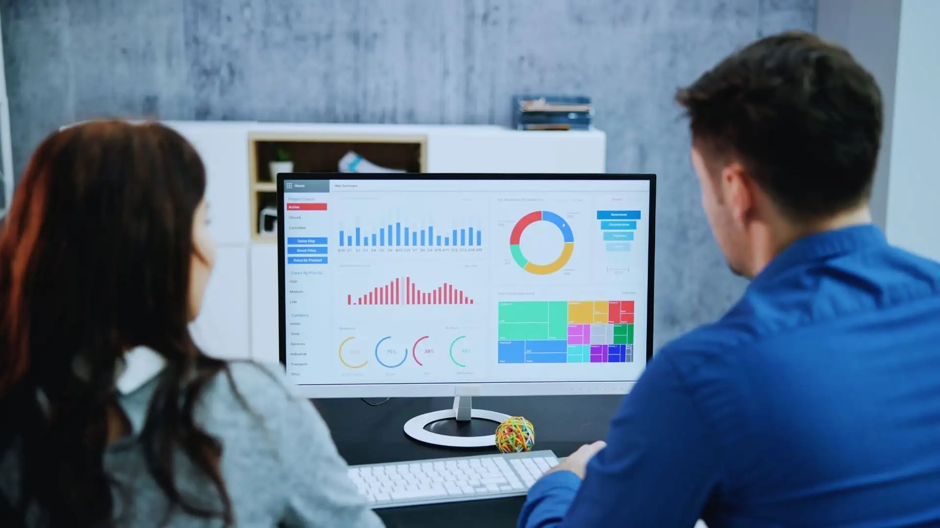 Two people sit at a desk in a modern office, reviewing colorful charts and graphs on their computer monitor as they discuss Monday.com price options and data visualizations.