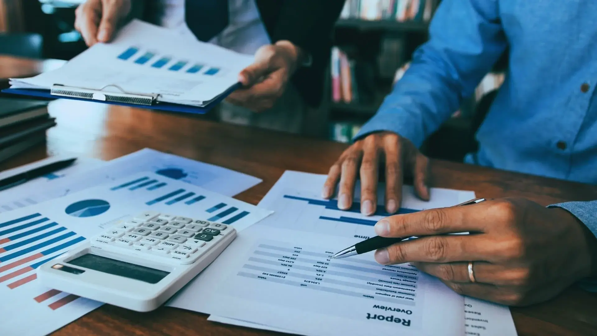 Two people reviewing printed graphs and financial reports at a desk, with a calculator and clipboard visible—perfect for comparing project management tools like TeamGantt vs Monday.com in a business or financial analysis setting.
