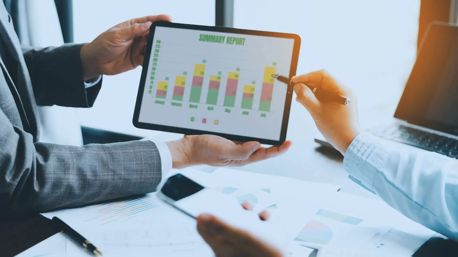 Two people in business attire review a tablet displaying a colorful bar chart summary report, possibly comparing Monday.com Price options. One points at the chart while the other holds the tablet; papers and a smartphone sit on the table.