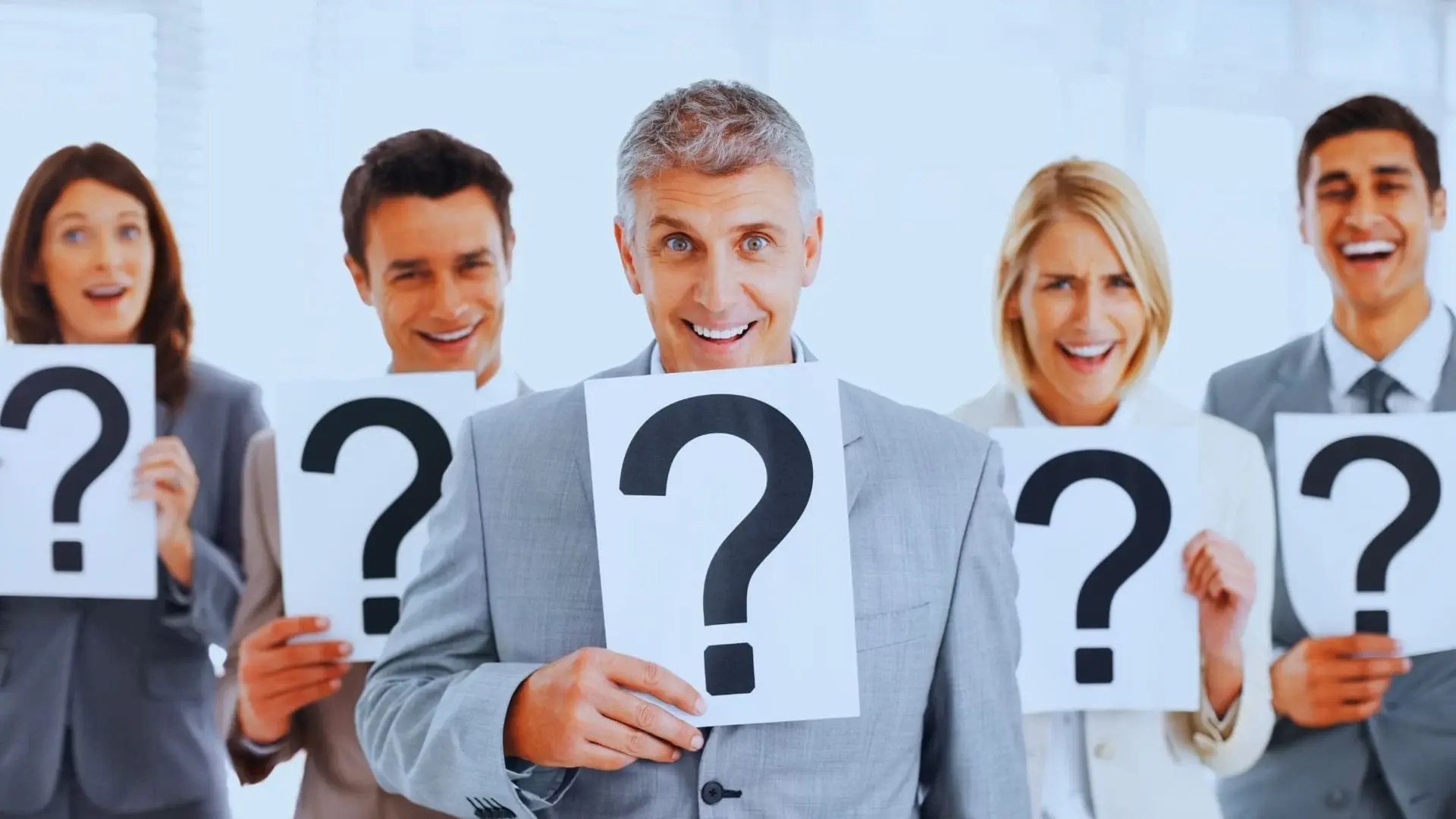 Four professionally dressed people stand in a row, smiling and holding large question mark signs in front of their faces, highlighting the dilemma of choosing between TeamGantt vs Monday.com in a bright, modern office setting.