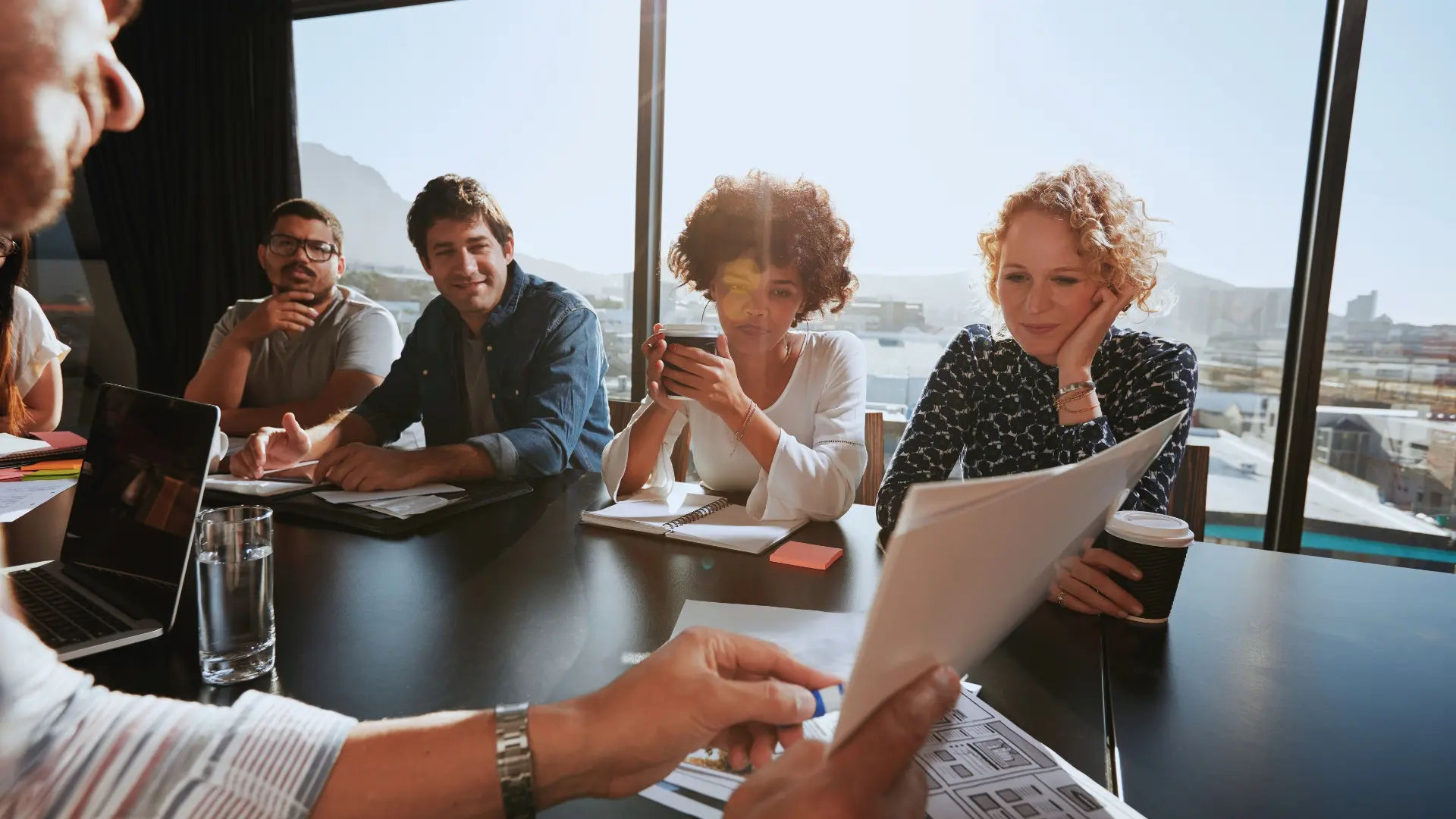 A group of four people sits at a conference table, attentively listening as someone hands out documents during a Planning in Agile session. Notebooks, coffee cups, and digital devices are on the table; large windows reveal a cityscape outside.