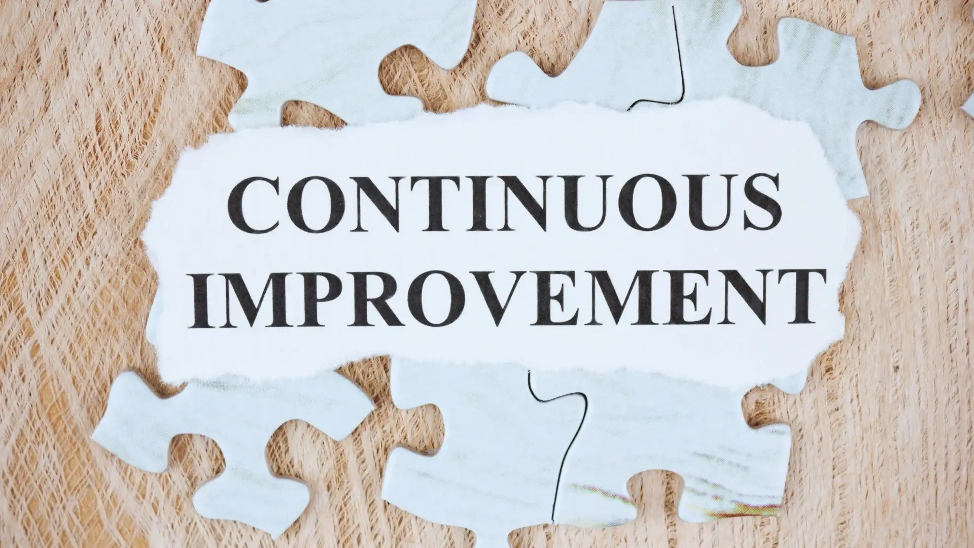 A torn piece of paper with the words "CONTINUOUS IMPROVEMENT" lies on top of light blue puzzle pieces, symbolizing planning in Agile, over a wooden surface.