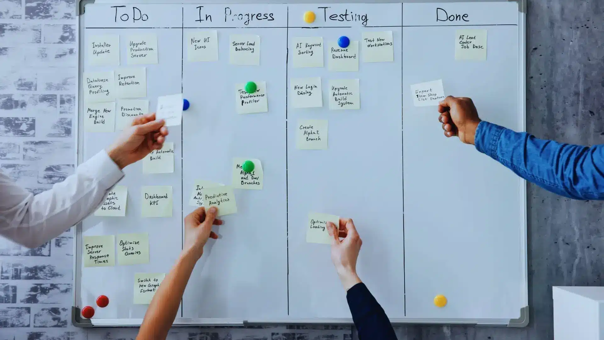 Four hands place sticky notes on a kanban board with columns labeled To Do, In Progress, Testing, and Done—demonstrating Agile Methodology definitions in action. The board is on a white wall and uses colorful magnets to attach notes.