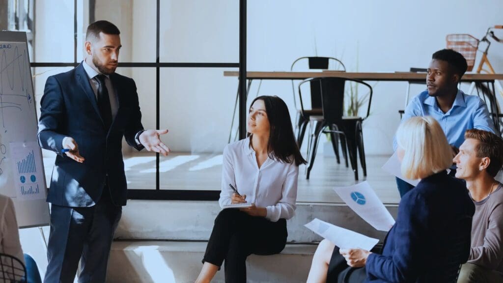 A man in a suit presents a graph on a flip chart to a group of colleagues seated and attentively listening in a modern office setting. One woman takes notes while others hold documents, engaging in an Agile Coaching session marked by collaborative discussion.