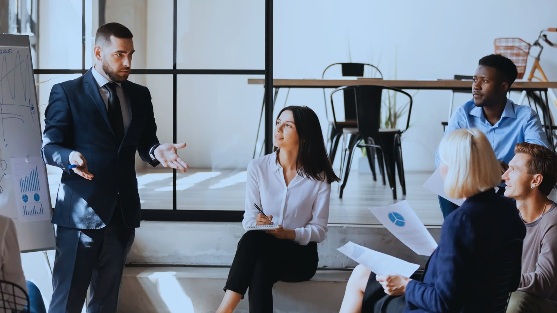 A man in a suit presents a graph on a flip chart to a group of colleagues seated and attentively listening in a modern office setting. One woman takes notes while others hold documents, engaging in an Agile Coaching session marked by collaborative discussion.