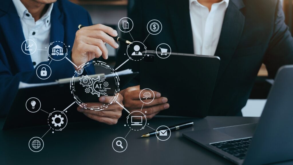 Two people in business suits discuss data at a desk with a tablet and laptop, overlaid with digital icons representing technology concepts like AI, cloud computing, and online security—highlighting key goals for change management.