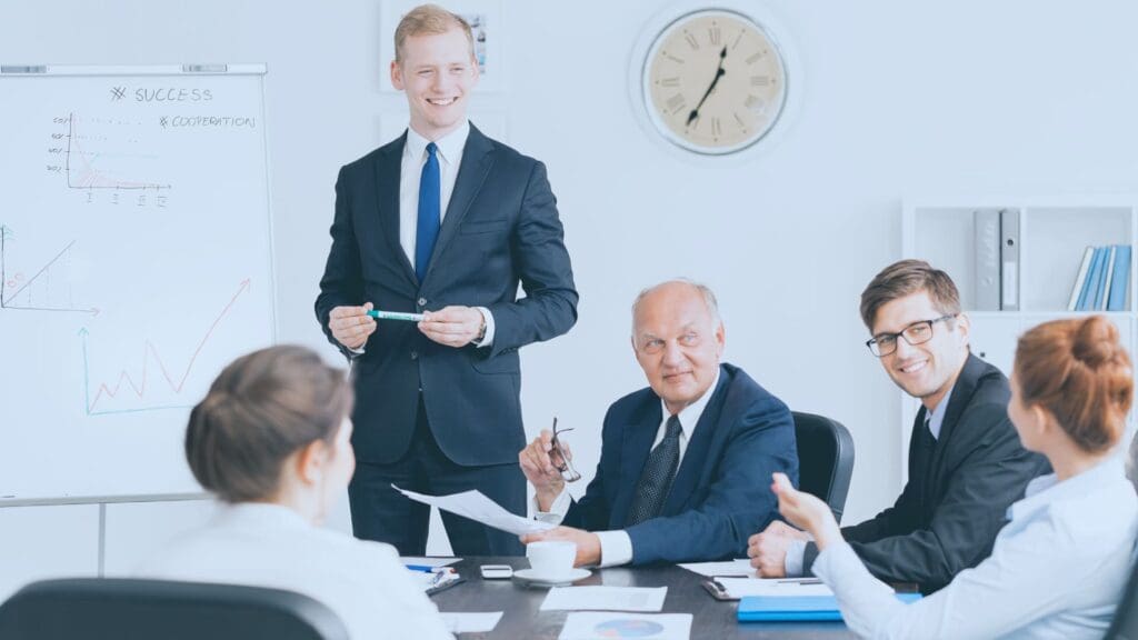 Five business professionals in a meeting room are engaged in discussion. A young man in a suit stands beside a flipchart with graphs related to Make Automations. Four colleagues, two men and two women, sit around a table, one man taking notes. A wall clock and shelves are in the background.