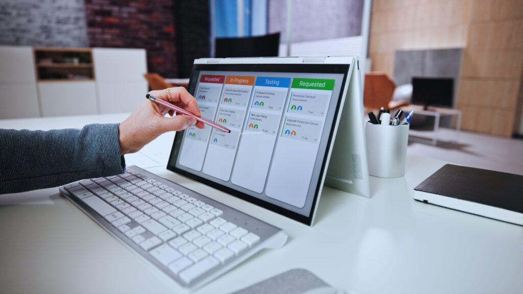 A person uses a stylus to interact with a tablet displaying a Gantt Chart for Agile Methodology, while a keyboard, mouse, and a cup of pens sit on the white desk in an office environment.