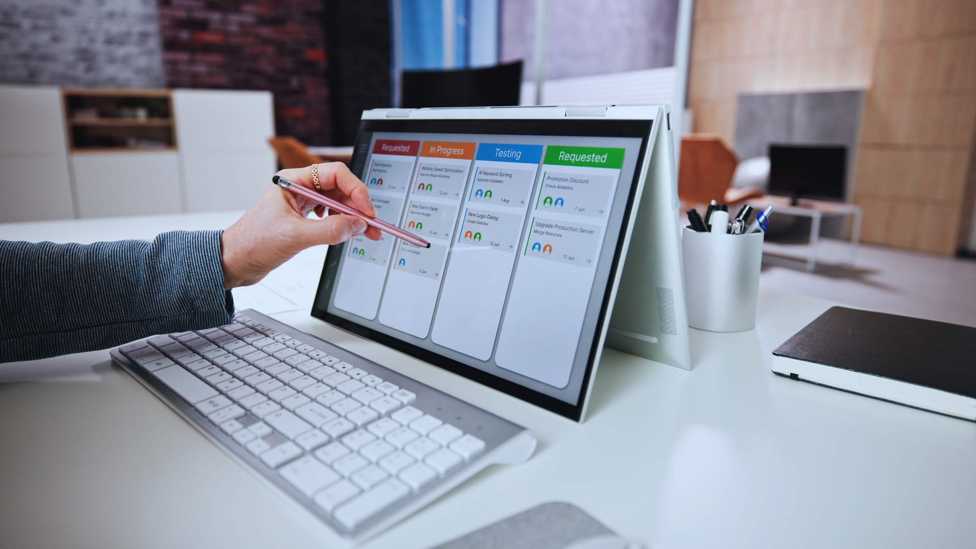 A person uses a stylus to interact with a tablet displaying a Gantt Chart for Agile Methodology, while a keyboard, mouse, and a cup of pens sit on the white desk in an office environment.