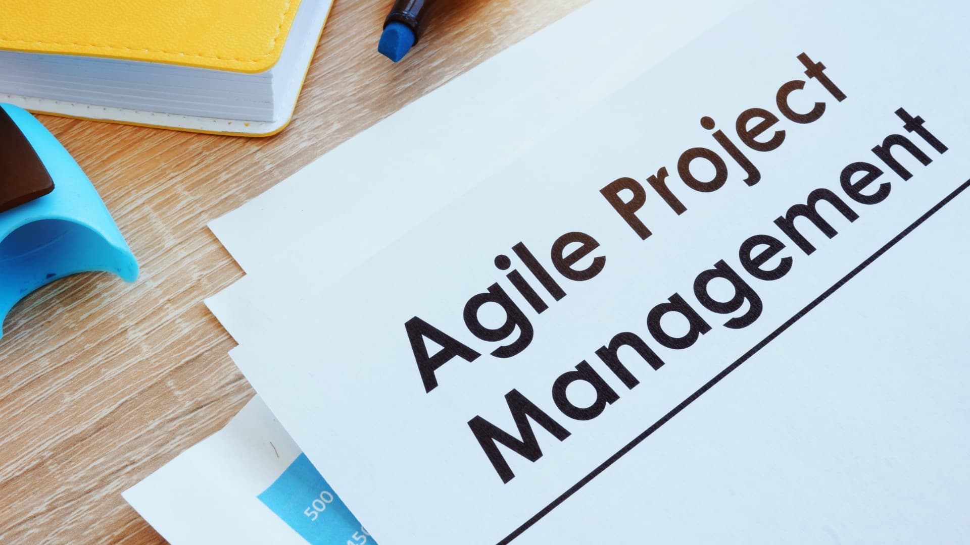 A document titled "Agile Project Management" sits on a wooden desk next to a yellow notebook, blue tape dispenser, and a pen—perfect for exploring planning in Agile methodologies.