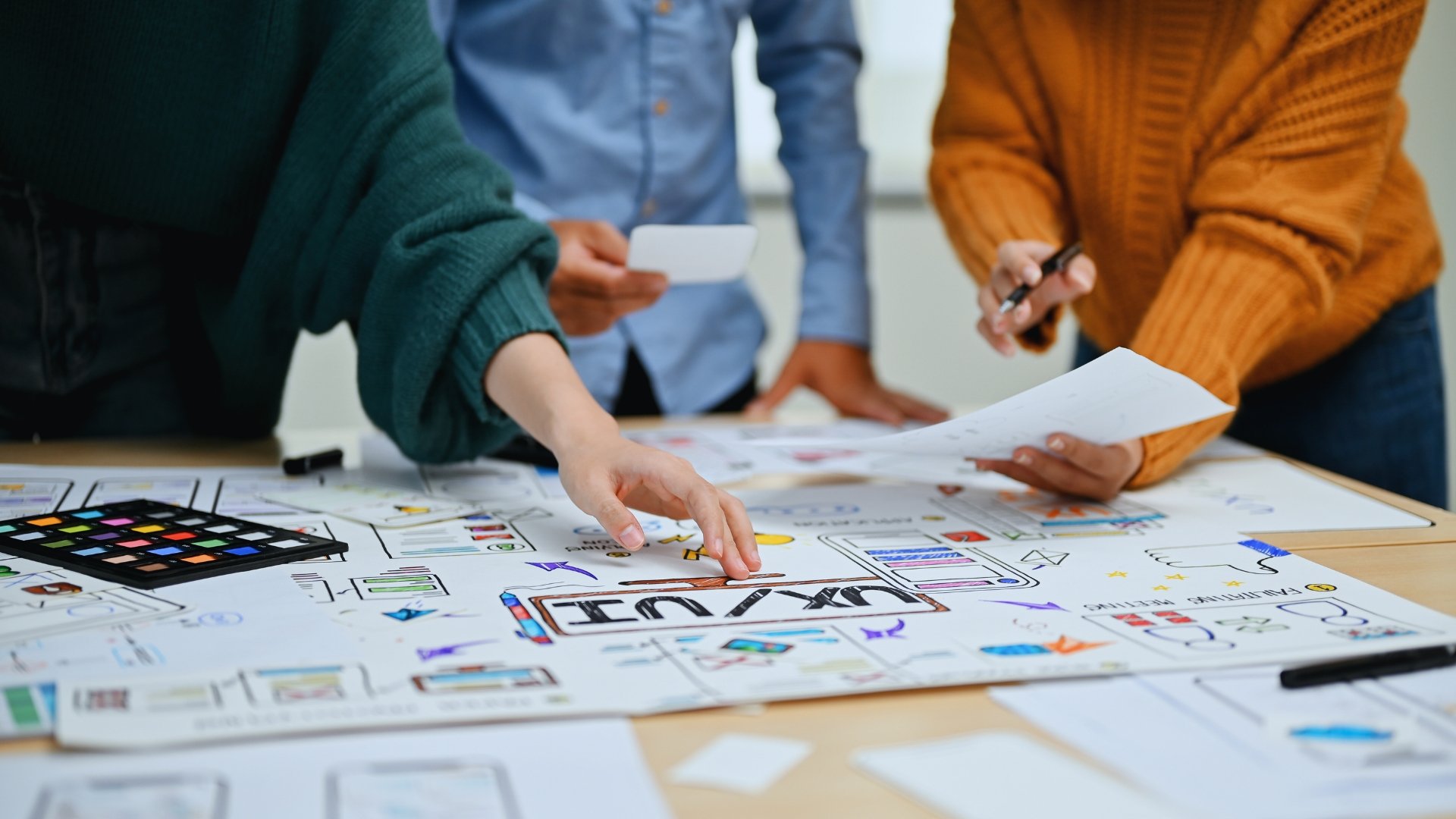 Three people collaborate at a table covered with UX/UI design sketches, paper prototypes, and notes, actively discussing and arranging design elements for a creative project within an Agile Programme framework.