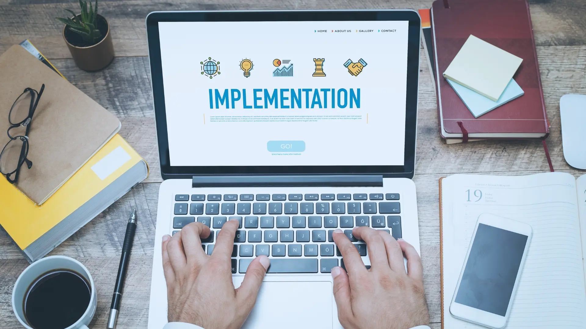 A person types on a laptop displaying the word "IMPLEMENTATION" alongside Agile Fundamental icons, with notebooks, papers, and a coffee cup scattered across a wooden desk.