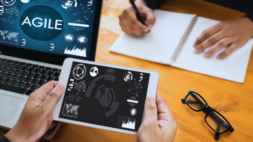 A person holds a tablet displaying digital graphics, while another writes in a notebook at a desk featuring a laptop with the word "AGILE," showcasing agile skills, along with glasses and scattered papers.