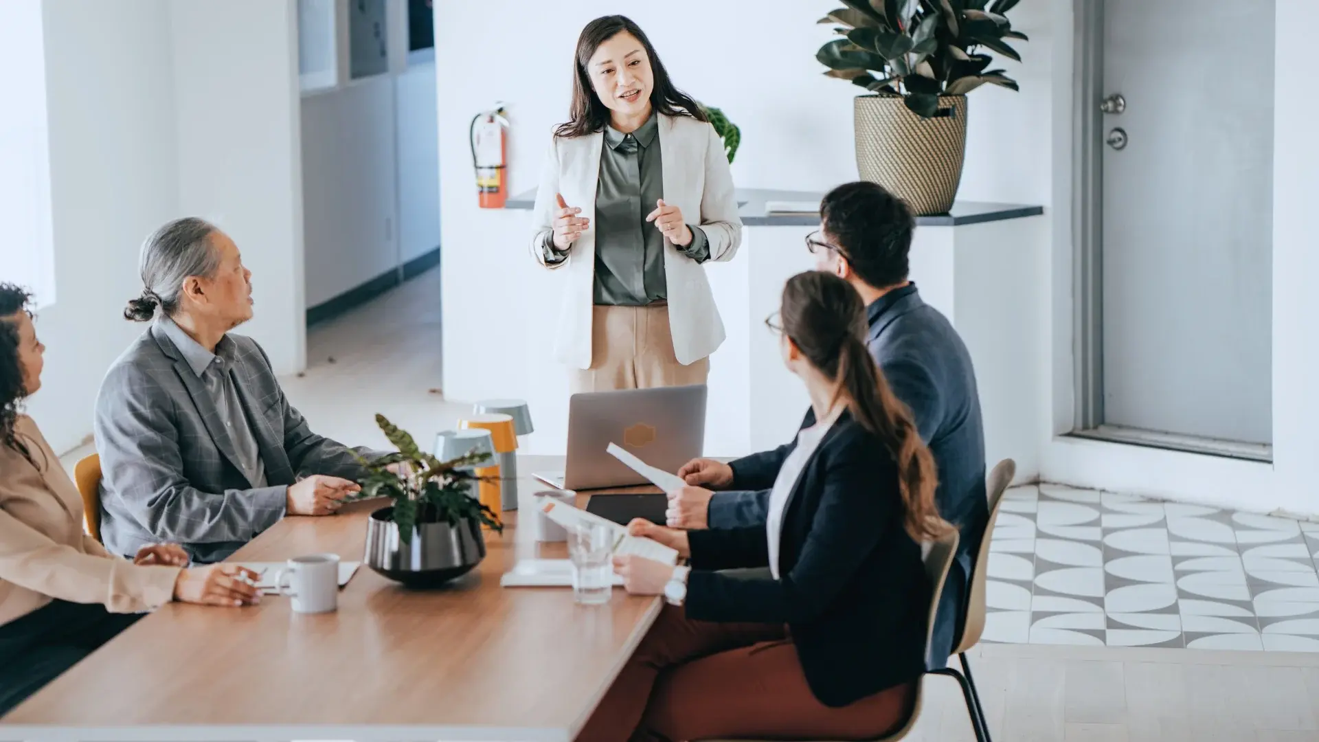 A woman stands and speaks to three seated colleagues during a meeting in a modern office, discussing PMI Stakeholder Analysis, with a laptop and documents on the table and a large potted plant in the background.