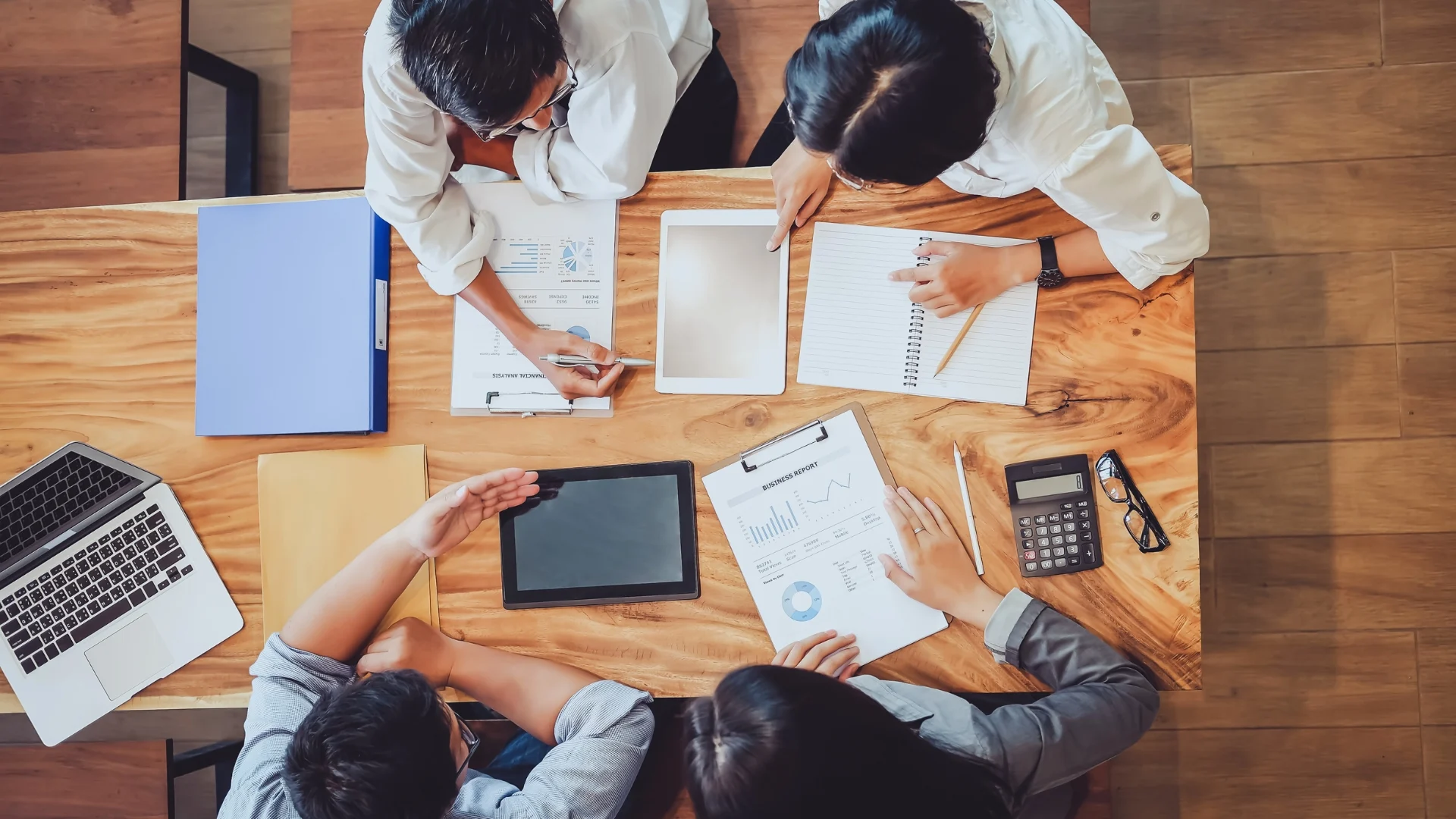 Four people sit around a wooden table, collaborating on documents, charts, and graphs. Tablets, a calculator, notebooks, and folders are spread out as they engage in Planning in Agile to organize their work.