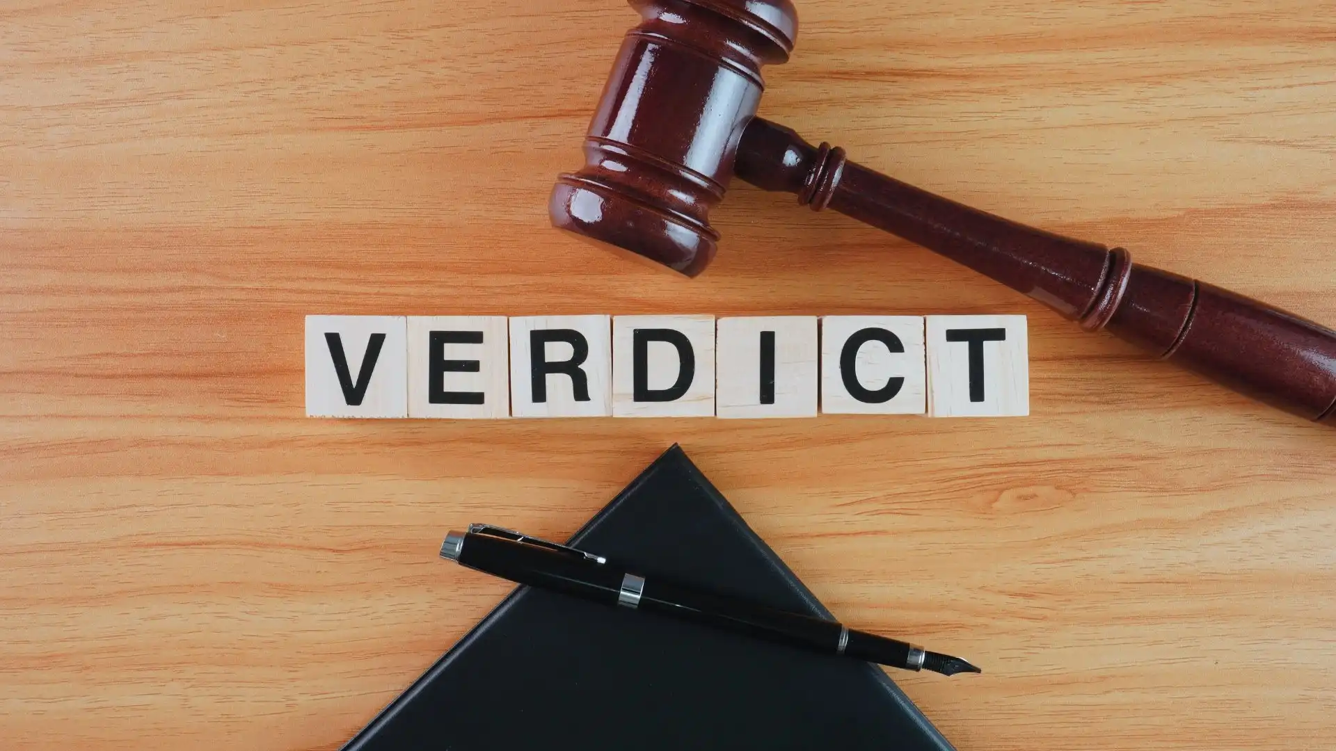 A wooden judge's gavel, letter blocks spelling "VERDICT," a black folder, and a pen are arranged on a light wooden surface—echoing the clarity and structure found in TeamGantt Pricing.