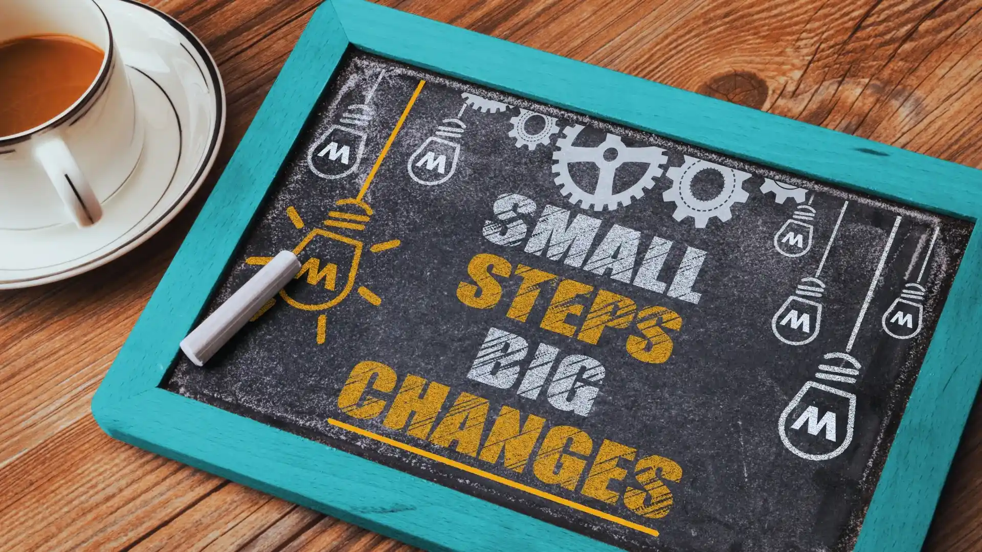 A turquoise-framed chalkboard on a wooden table displays "Small Steps Big Changes" in bold yellow and blue, symbolizing goals for change management, with chalk drawings of light bulbs and gears. A cup of tea sits nearby.