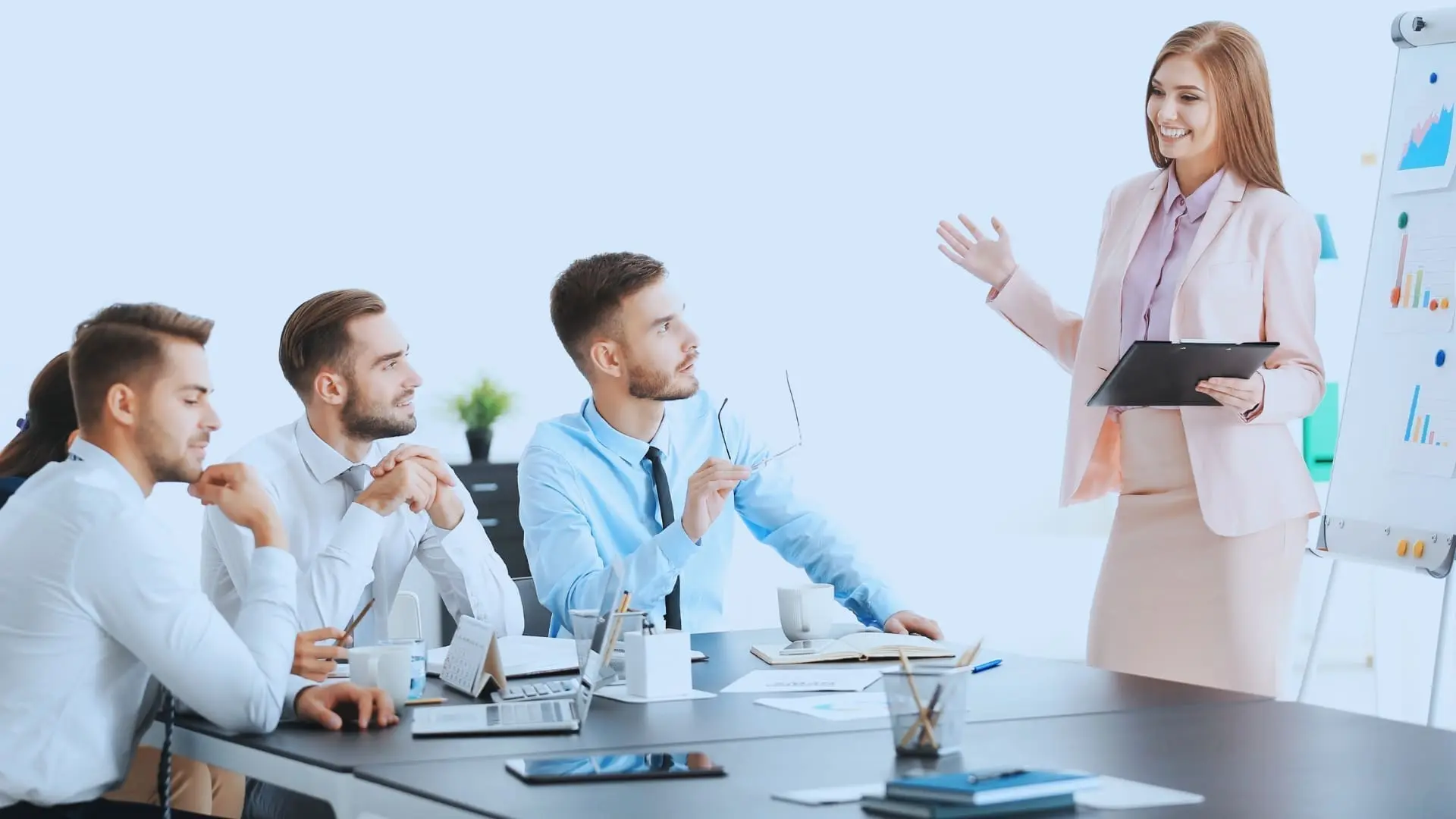 A woman in business attire stands and presents Agile Methodology definitions on a flipboard to four colleagues seated at a conference table, who are listening attentively. Laptops, papers, and coffee cups are on the table.