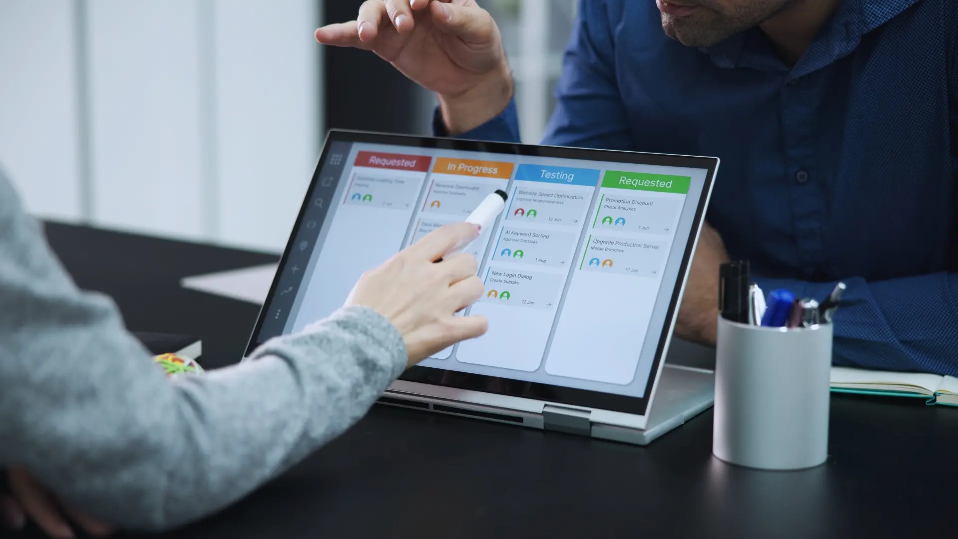 Two people discuss a project management dashboard on a convertible laptop, with one pointing at tasks labeled "Requested," "In Progress," and "Testing." A Gantt Chart for Agile Methodology is visible on the screen. A pen holder sits nearby on the desk.