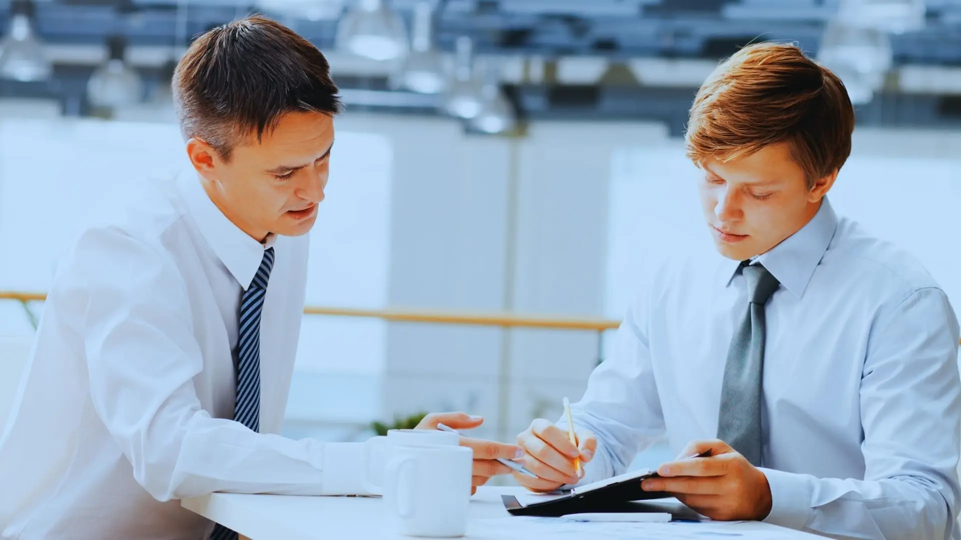 Two men in business attire sit at a desk, discussing documents. One points at a tablet with a pen while explaining Agile Coaching, as the other listens attentively. Coffee mugs and a modern office backdrop complete the scene.