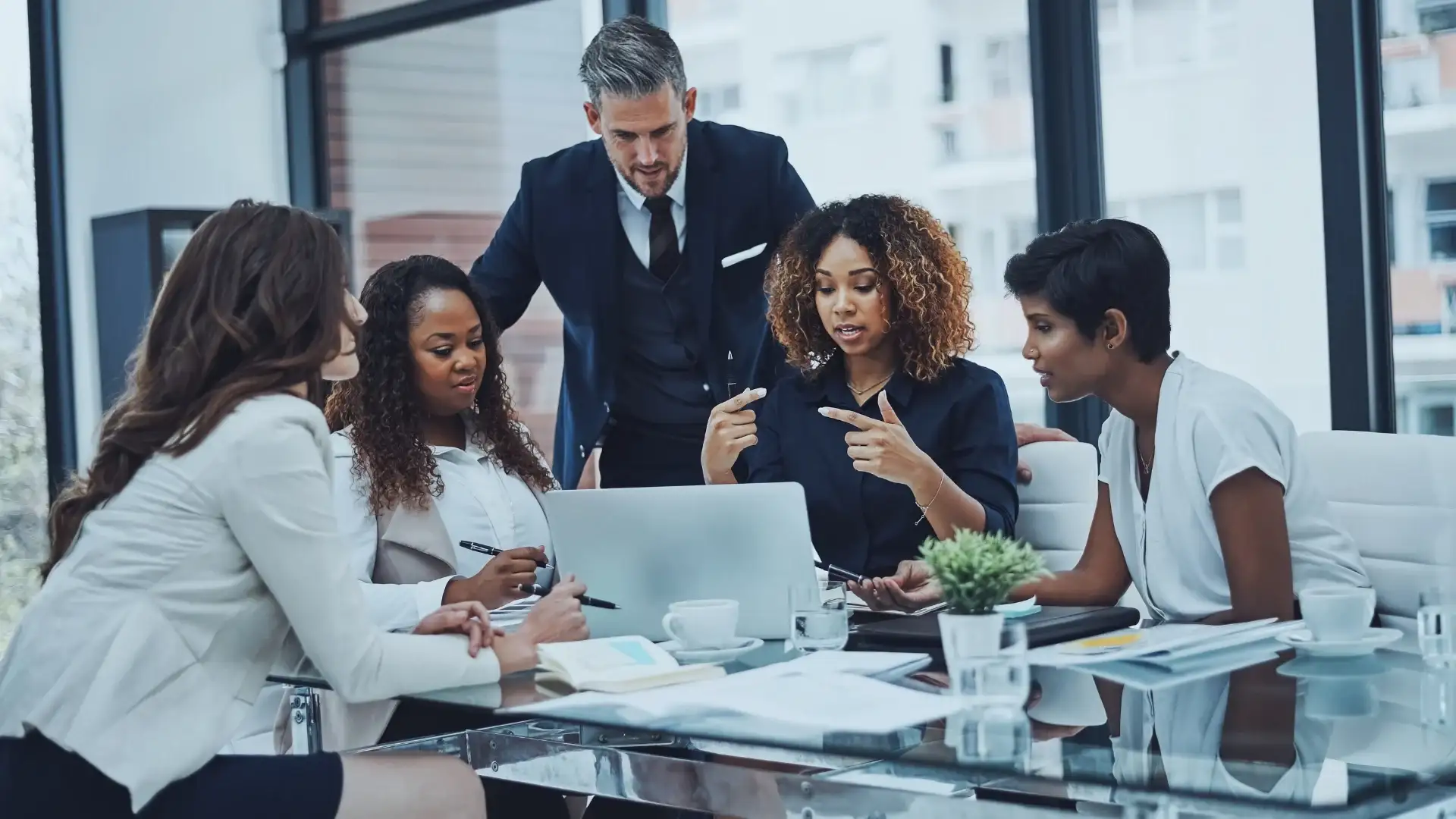 Five professionals, three women seated and one standing man, discuss Goals for Change Management around a glass table with documents, laptops, and a plant in a bright office with large windows.