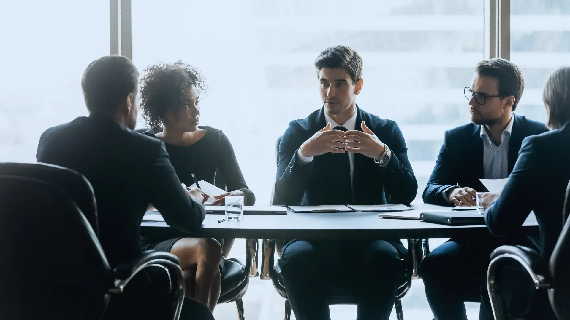 Four professionally dressed people sit around a table in a bright office, engaged in a serious business discussion about what is an agile environment, with papers and laptops in front of them.