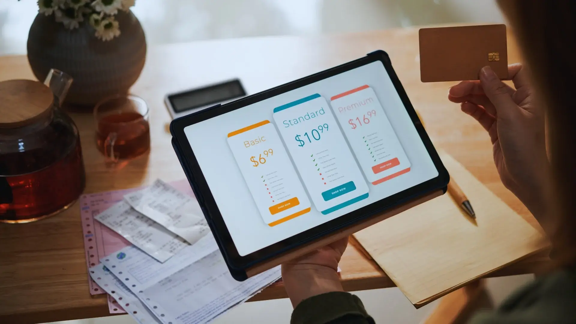 A person holds a tablet displaying subscription plan options—Basic, Standard, and Premium with Monday.com fees—while holding a credit card. Papers, a pen, tea, and a phone are on the wooden table in the background.