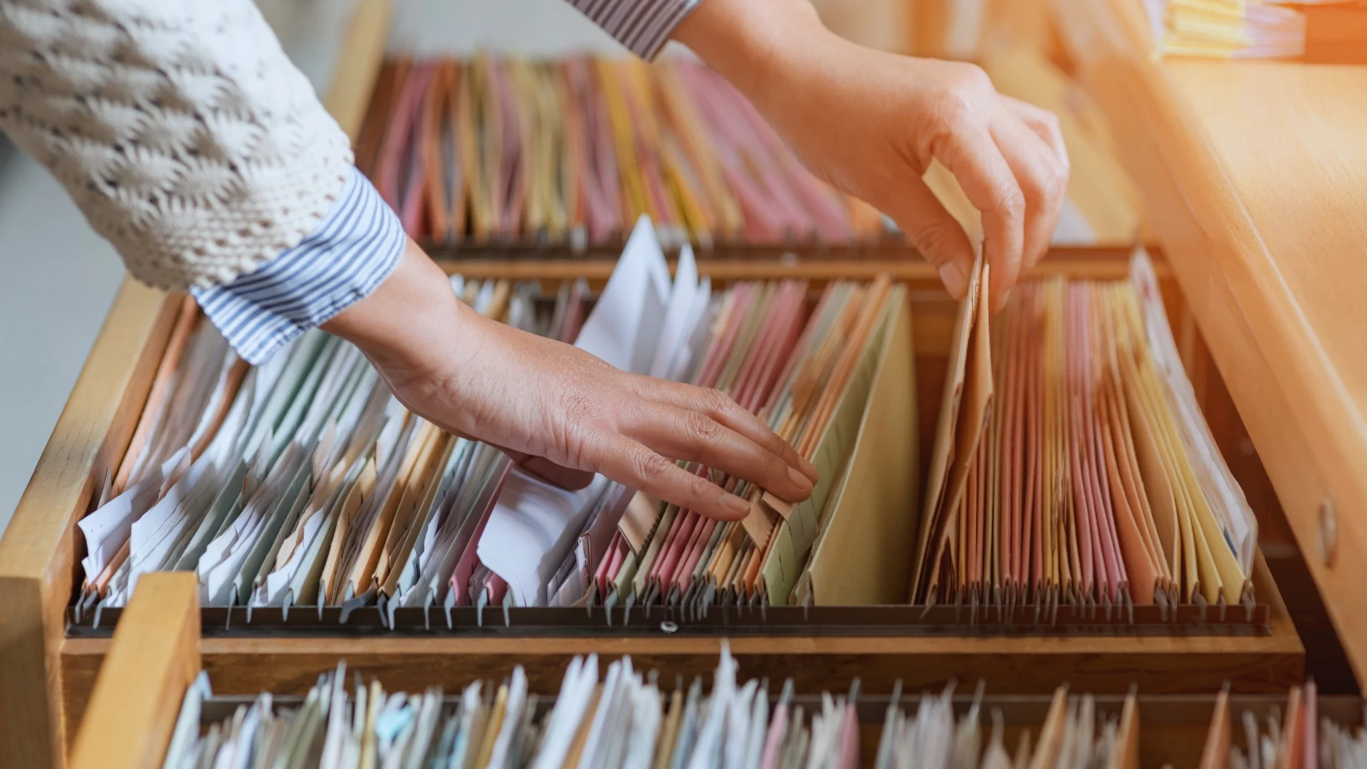 Close-up of a person’s hands searching through files in a wooden filing cabinet drawer, with labeled tabs visible—perfect for organizing documents like a Project Closure Email.