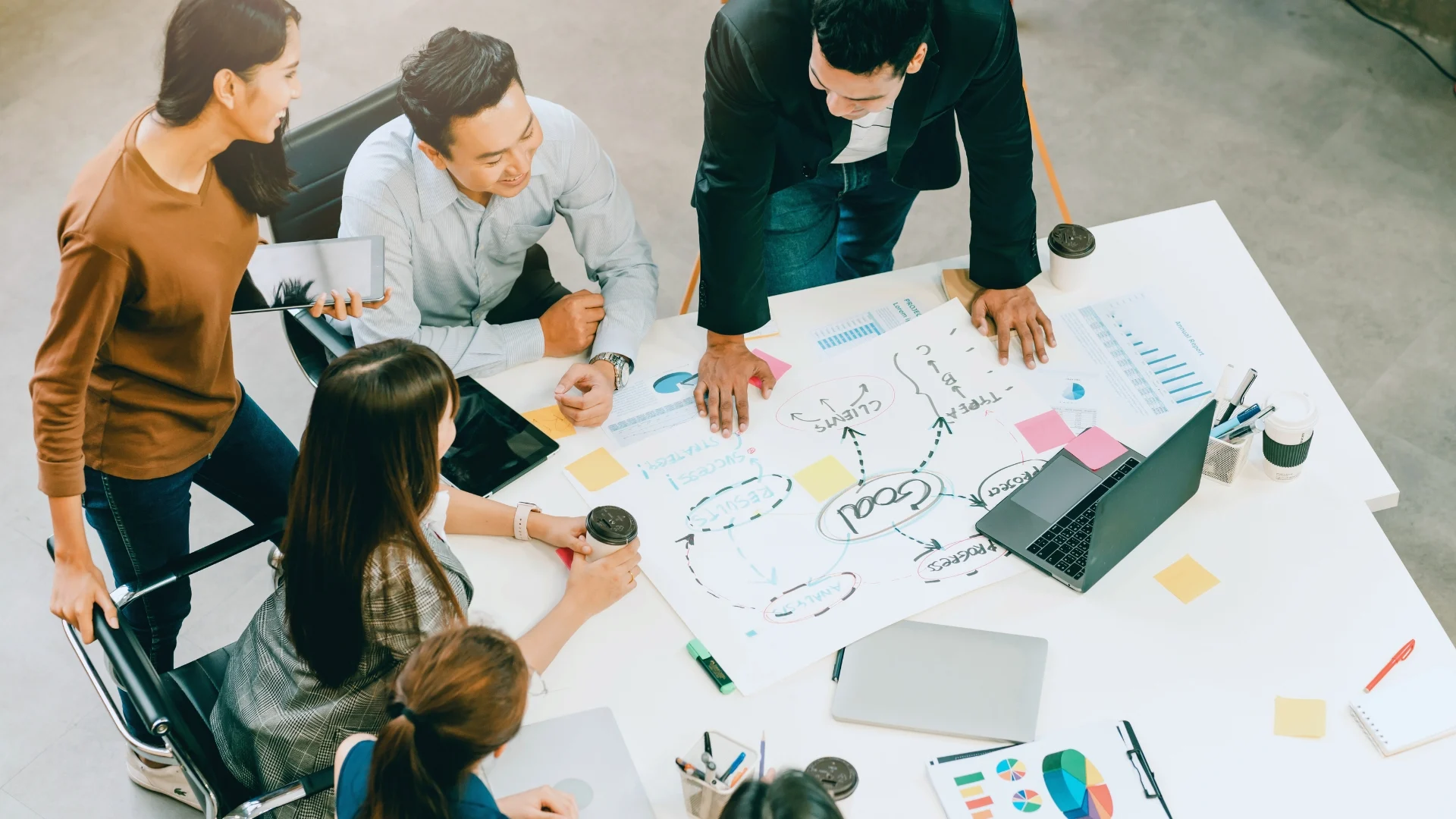 A group of five people collaborate around a table with charts, a laptop, coffee cups, and brainstorming notes, focusing on value-driven delivery in a bright office setting, viewed from above.
