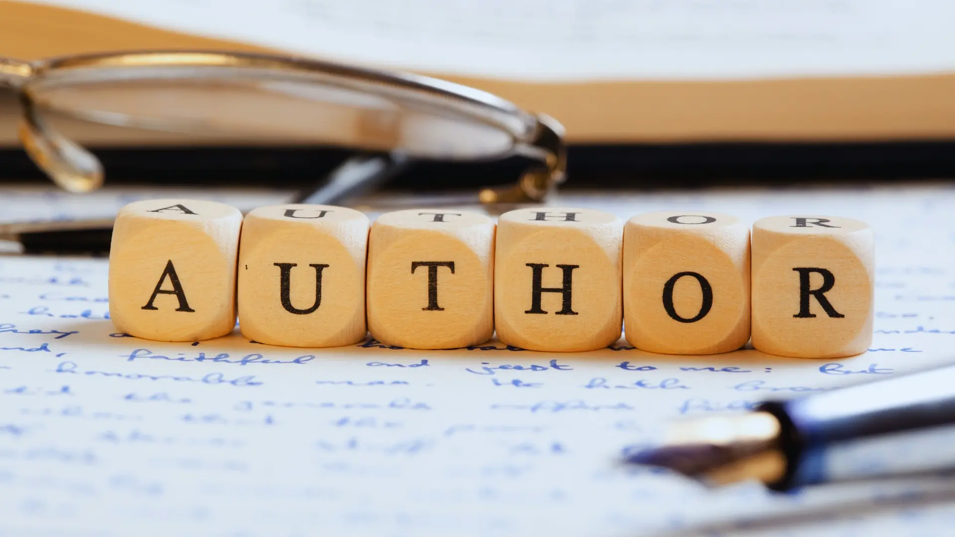 Close-up of wooden letter blocks spelling "AUTHOR" on handwritten papers, with eyeglasses and an open book in the background, a pen resting nearby—discover a creative BVDash Alternative for your writing journey.
