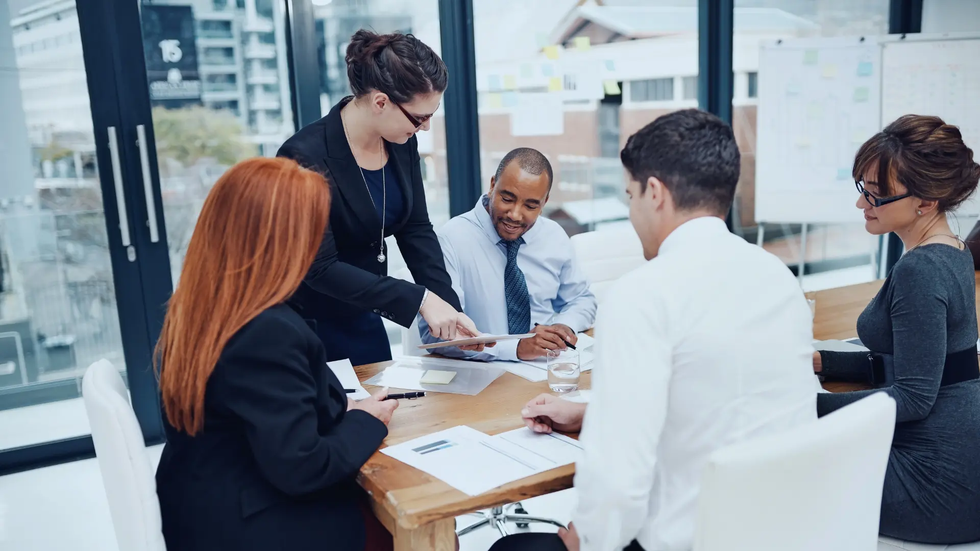 Four professionals sit around a wooden table in a modern office with large windows. As they discuss project details and Monday.com fees, a woman stands, pointing to a document, while the others listen intently. Papers and glasses of water are on the table.
