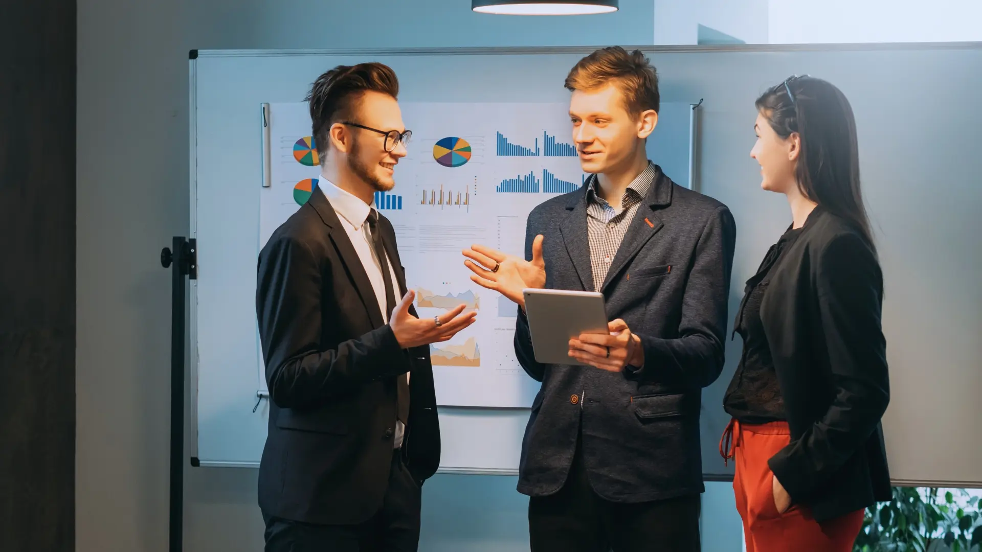 Three people in business attire stand and talk in front of a whiteboard displaying colorful charts and graphs, illustrating what is an agile environment. One holds a tablet as they engage in a dynamic discussion.