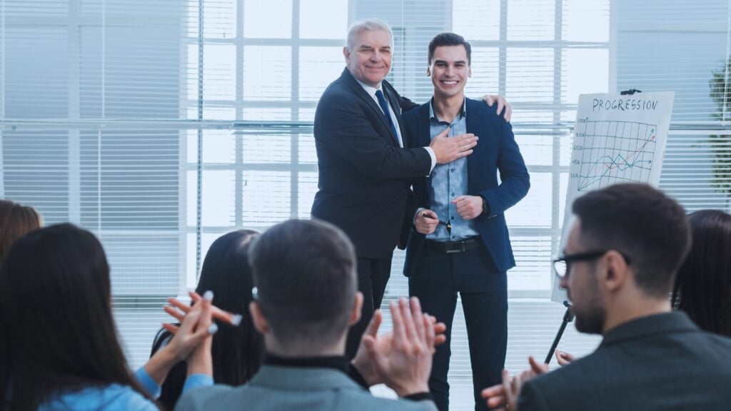 Two men in suits smile and stand together at the front of a room, while an audience applauds them. A flip chart labeled "Progression" with graphs details Agile Project Examples. The setting appears to be a business meeting or presentation.