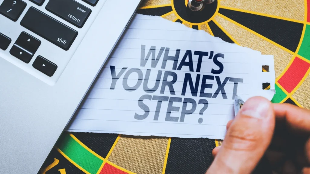 A hand writes on a notepad with the words “WHAT’S YOUR NEXT STEP?” placed on a dartboard next to a laptop keyboard, hinting at decision-making or planning in what is an agile environment.
