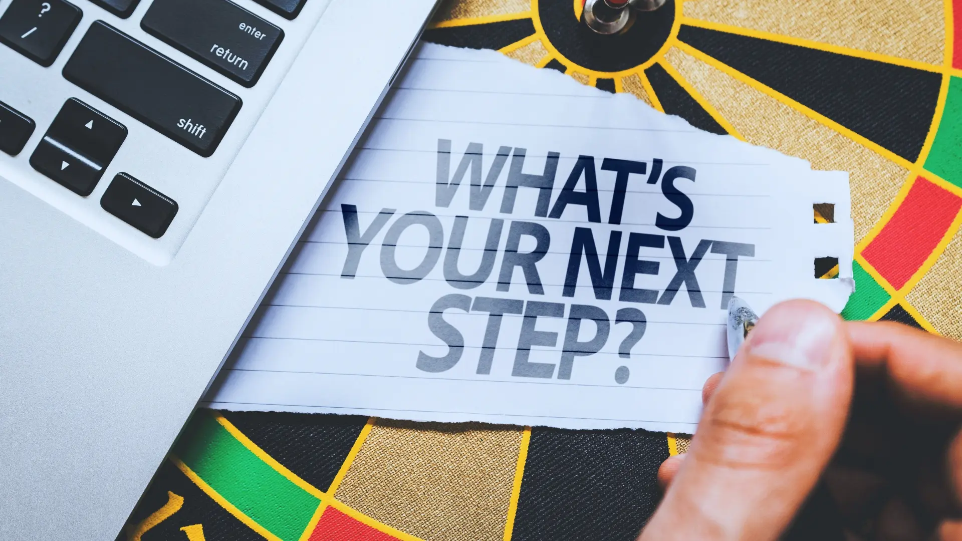 A hand writes on a notepad with the words “WHAT’S YOUR NEXT STEP?” placed on a dartboard next to a laptop keyboard, hinting at decision-making or planning in what is an agile environment.