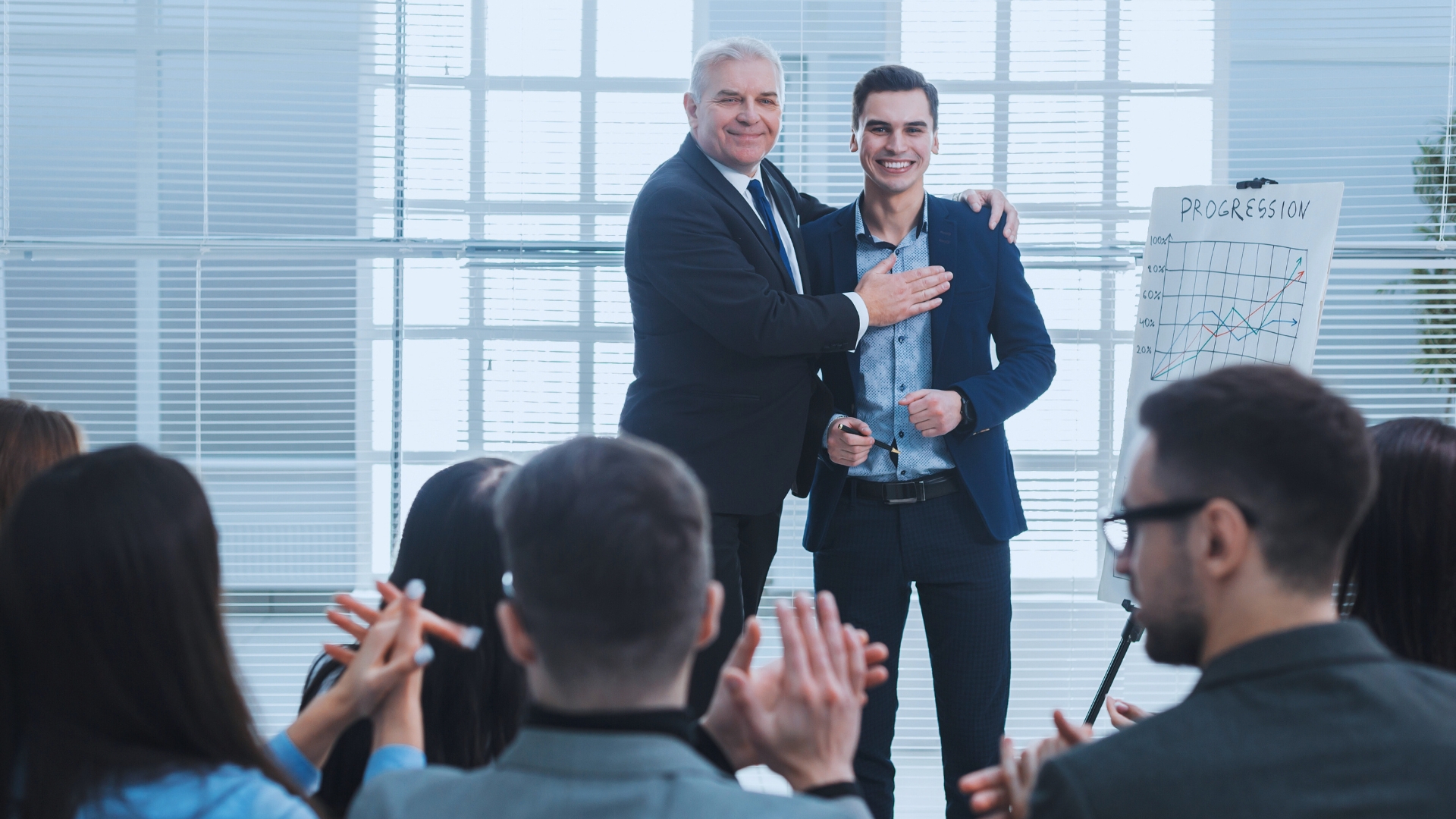 Two men in suits smile and stand together at the front of a room, while an audience applauds them. A flip chart labeled "Progression" with graphs details Agile Project Examples. The setting appears to be a business meeting or presentation.