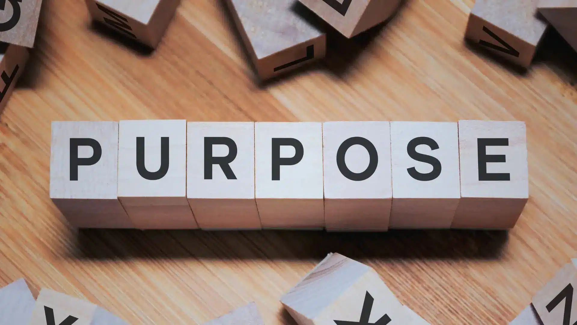 Wooden blocks arranged in a row spell out "PURPOSE" in bold black letters, symbolizing Project Management Innovations, with additional scattered wooden blocks on a wooden surface.