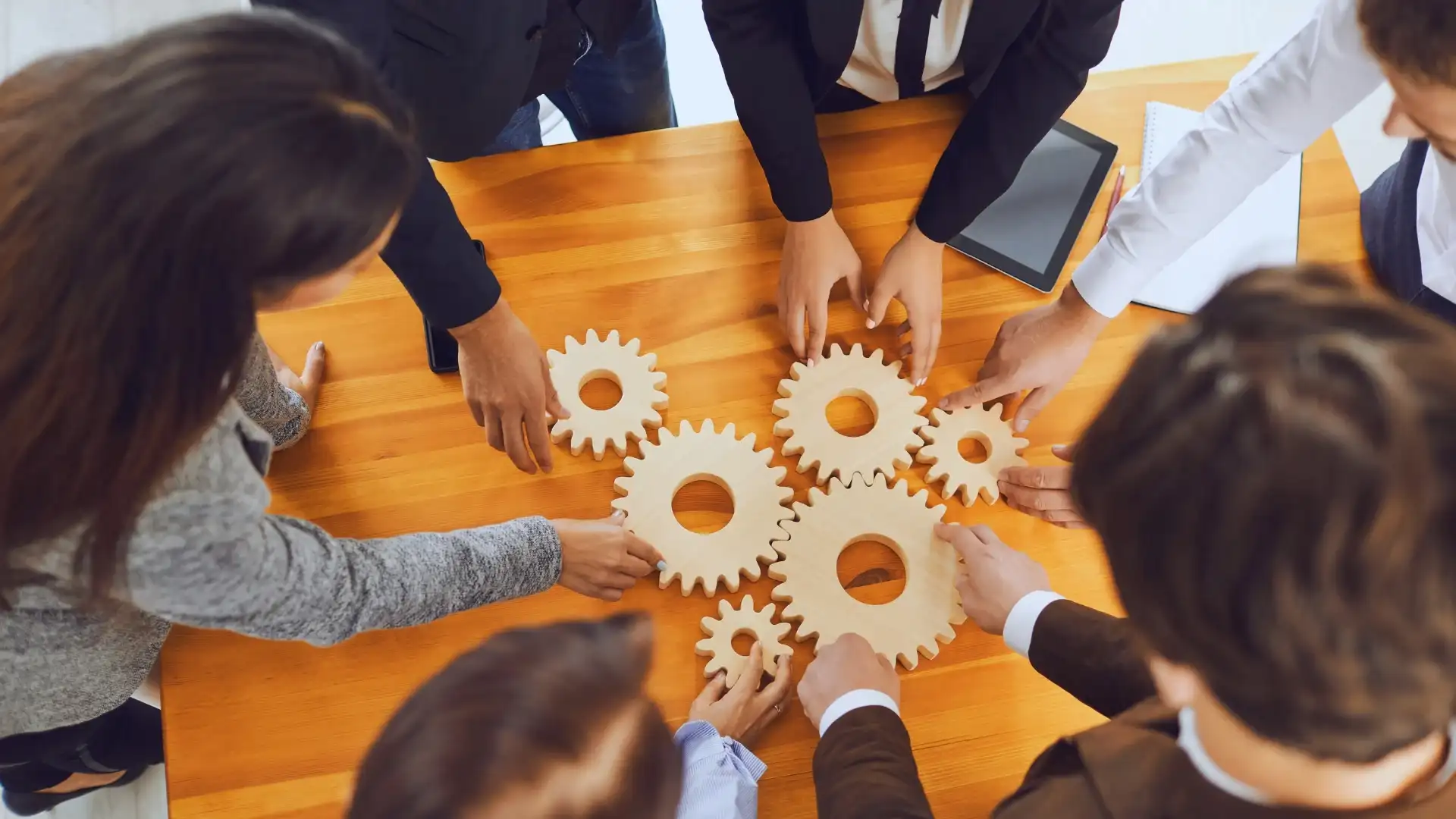 A group of people standing around a wooden table fitting wooden gears together, symbolizing teamwork and collaboration—a nod to the functions of project management office. A tablet is placed on the table.