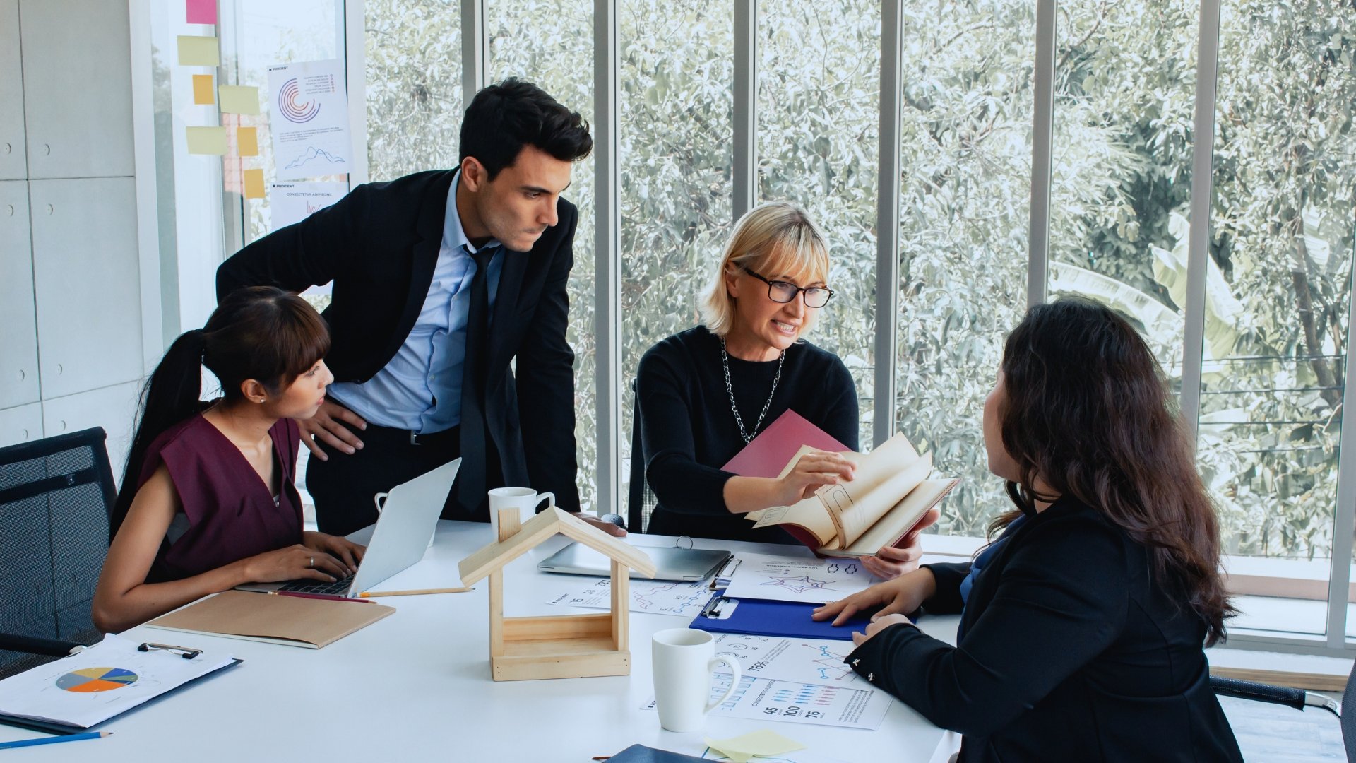 Four colleagues in business attire discuss On-Time Project Management around a table with documents, charts, a laptop, and a small wooden house model, in a bright office with large windows and trees outside.