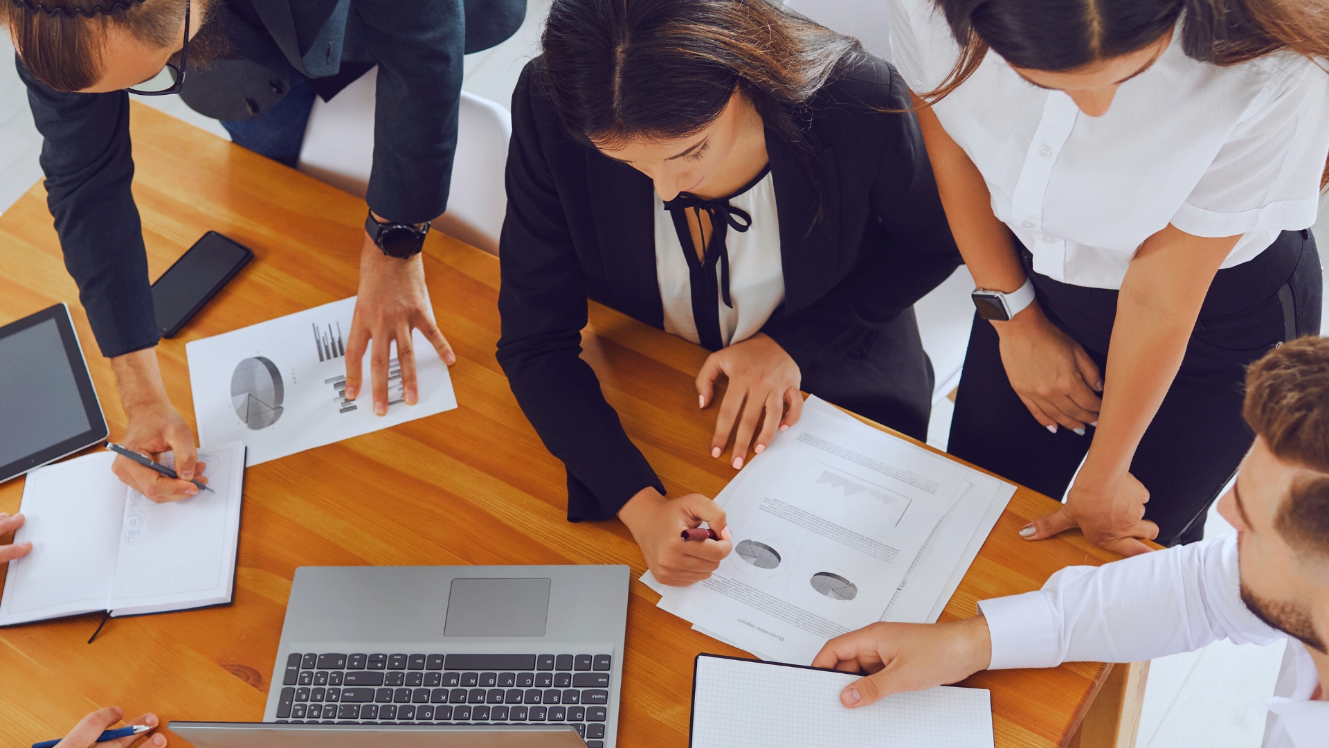 Five people gather around a wooden table, reviewing charts and graphs, taking notes, and discussing the functions of project management office during a collaborative work session.