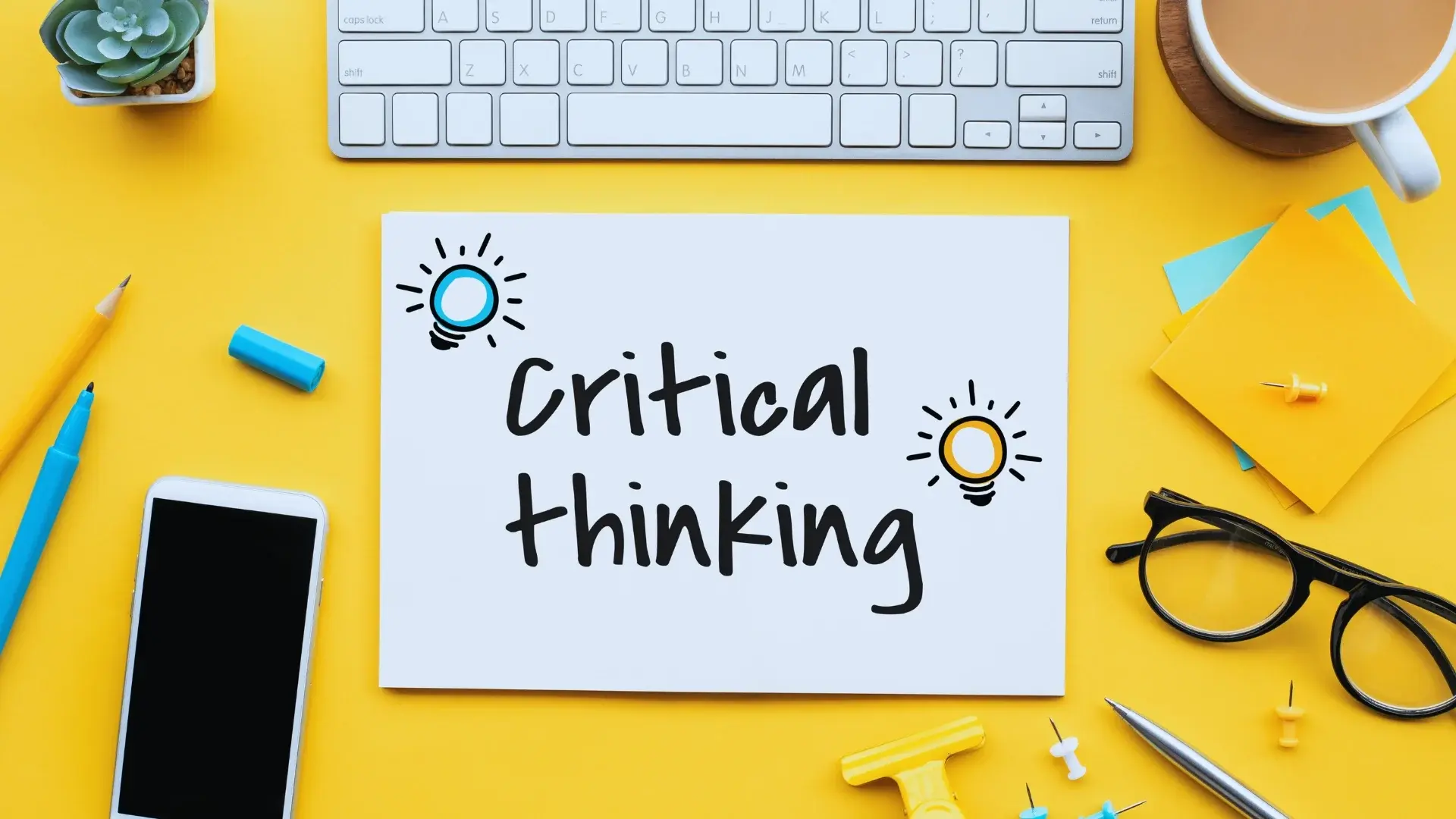 A white paper with "Critical thinking" written on it, decorated with blue and yellow lightbulb doodles, sits on a yellow desk surrounded by a keyboard, smartphone, pen, pencil, stationery items, and an On-Time Project Management guide.