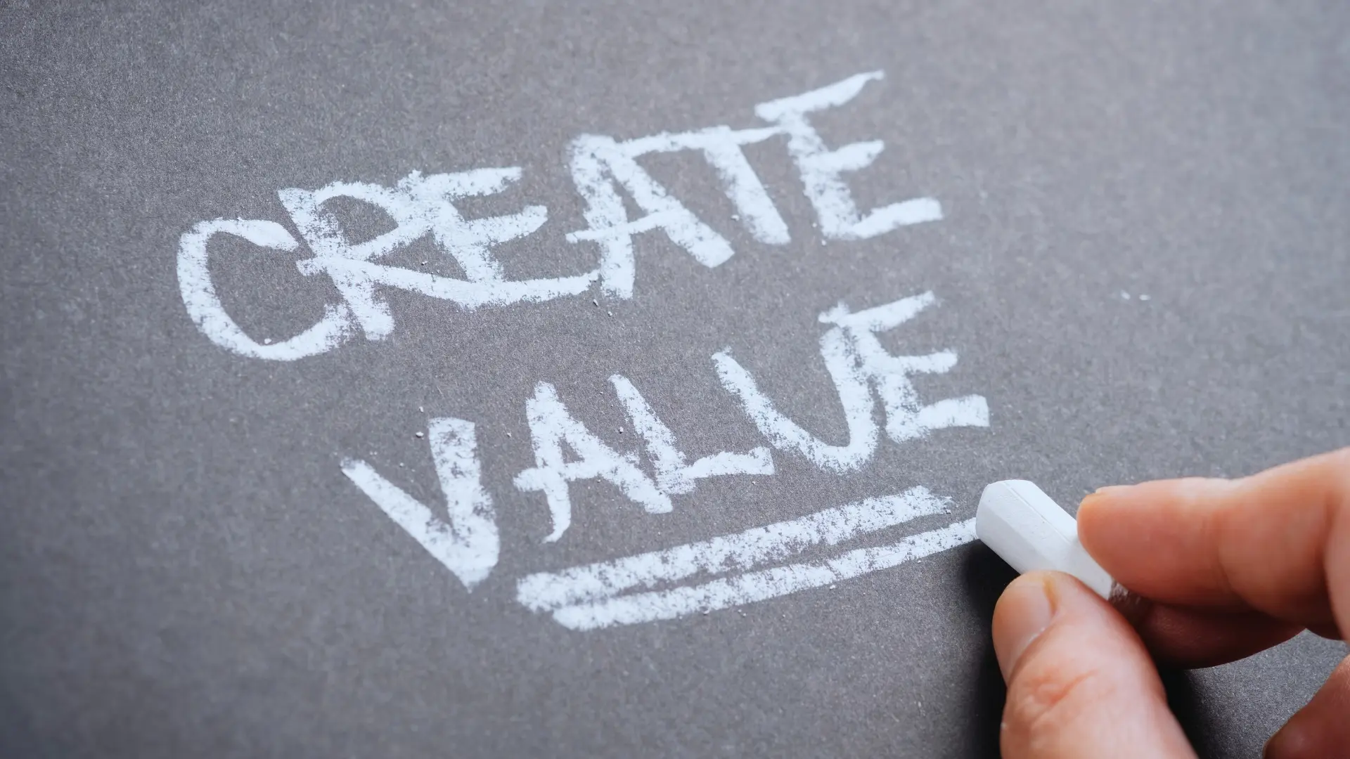 A hand writes "CREATE VALUE" in large, white capital letters with chalk on a dark surface, highlighting the importance of Value-Driven Delivery by underlining the word "VALUE.