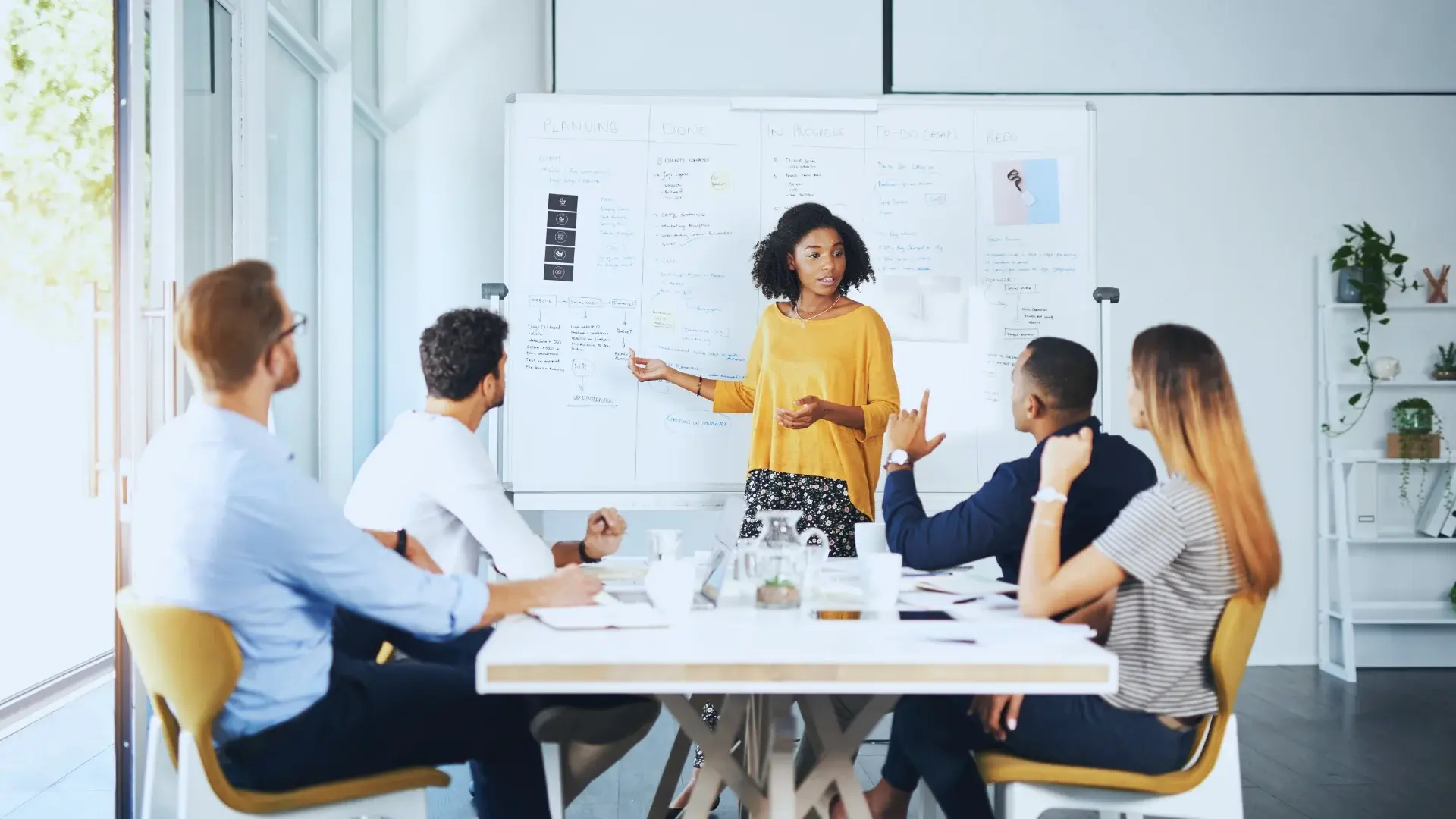A woman stands and presents information on a whiteboard to four colleagues seated around a conference table, engaging in a business meeting in a bright, modern office focused on the functions of project management office.