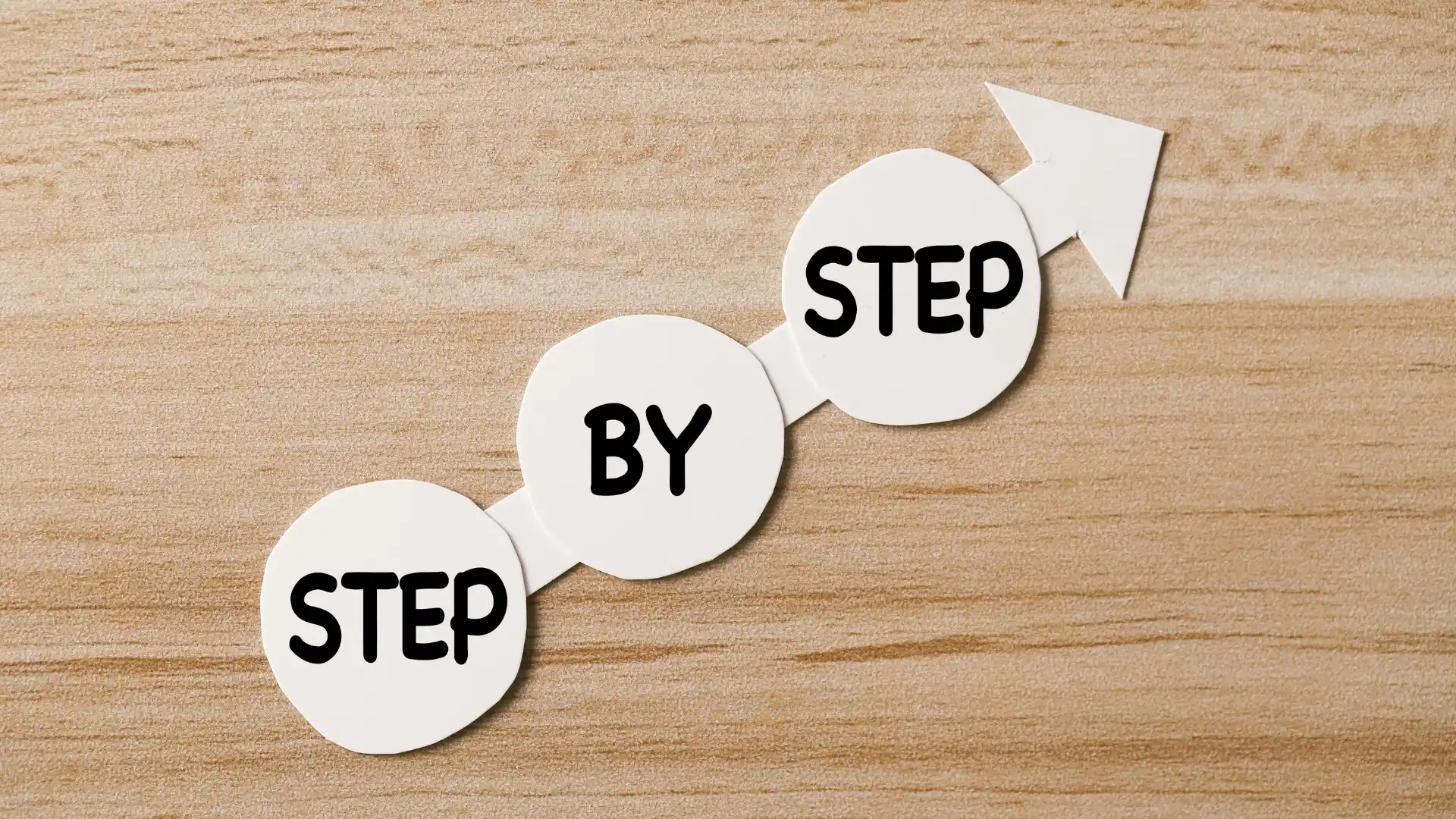 Three white circles with the words "STEP BY STEP" are aligned diagonally on a wooden surface, connected by a white arrow pointing upward to the right, symbolizing progress—much like comparing SmartSuite vs Taskade for productivity.