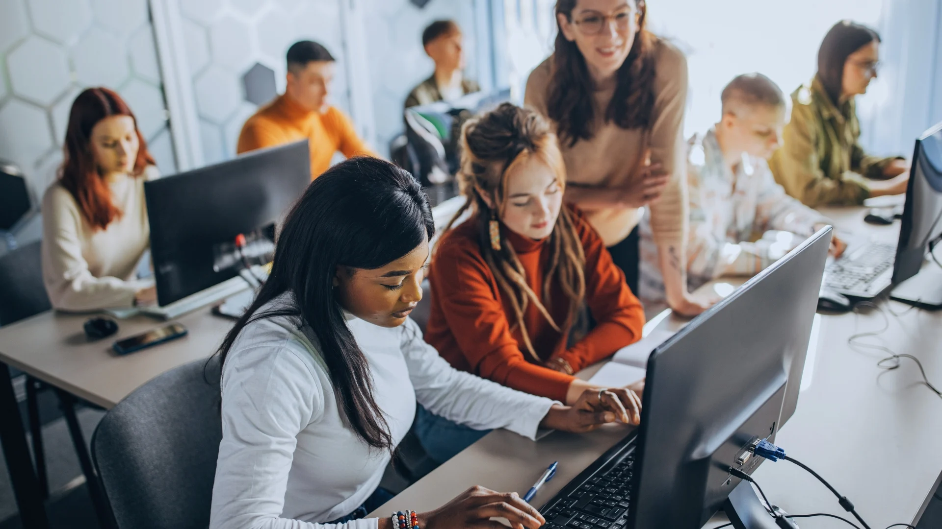 A diverse group of young adults works together at computers in a modern classroom. Two women in the foreground focus on a monitor while a teacher stands nearby, guiding students through Project Management Innovations.
