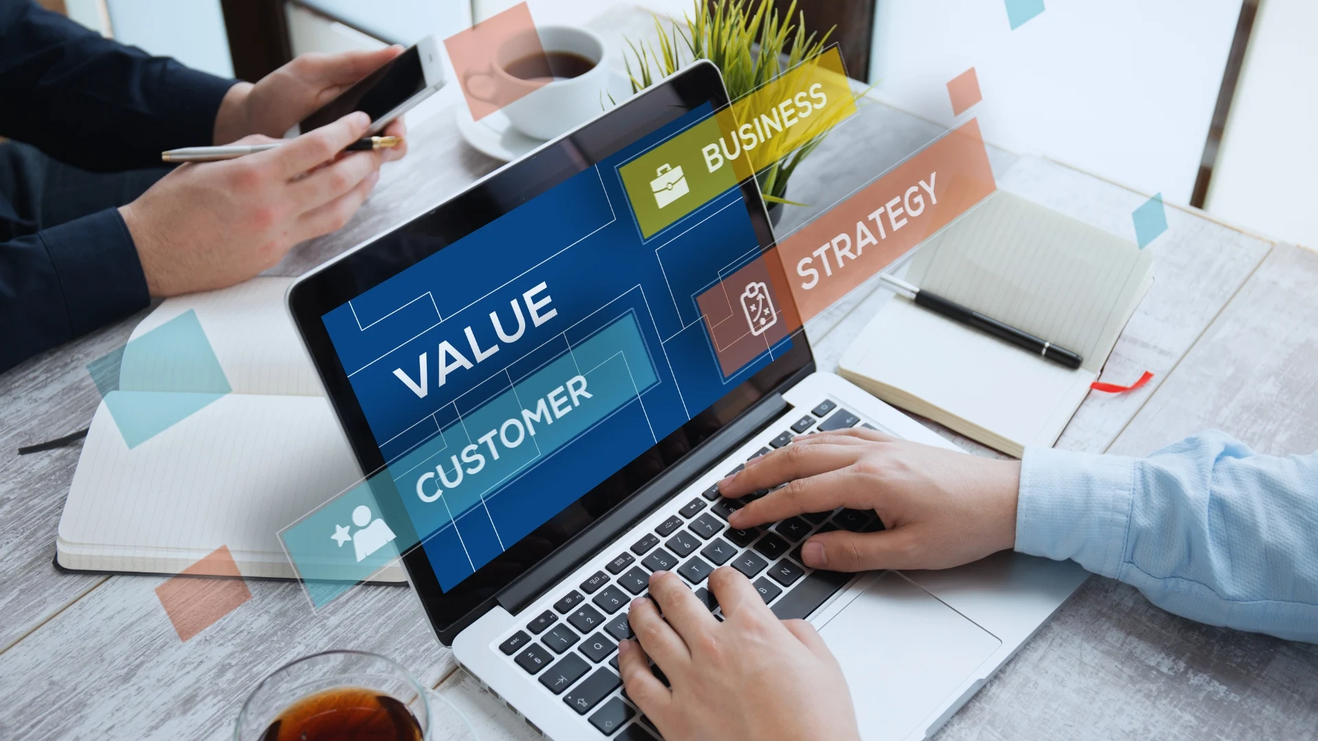 Person typing on a laptop displaying the word “VALUE,” with digital overlays showing “CUSTOMER,” “BUSINESS,” and “STRATEGY”—all reflecting a value-driven delivery approach. Another person uses a phone; notebooks, coffee, and plants rest on the table.