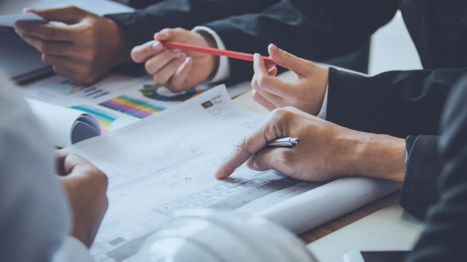 Close-up of several people’s hands discussing documents with charts and graphs. One person points at a paper with a pen, while another holds a red pencil, suggesting a collaborative business meeting or Project Closure Email planning session.