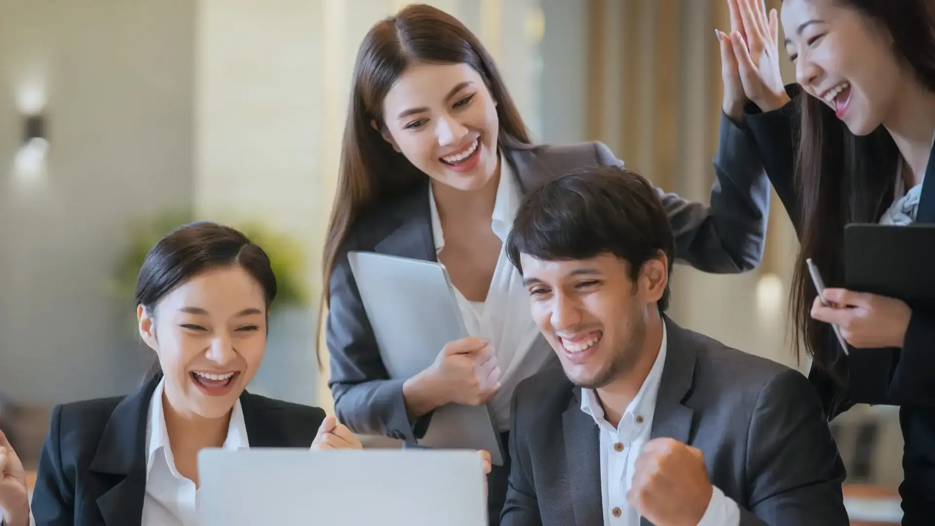 Four young professionals in business attire celebrate a successful project closure email, gathered around a laptop, smiling and cheering enthusiastically in a modern office setting.