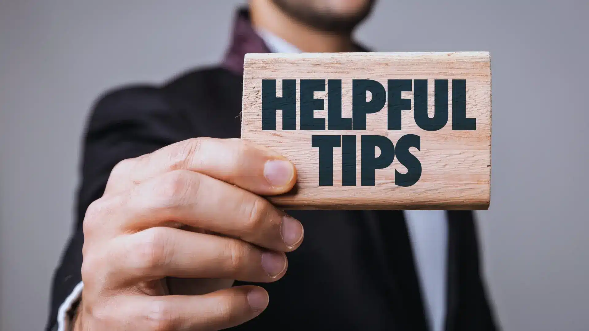 A person in a suit holds a wooden card towards the camera with the words "HELPFUL TIPS" printed in bold, black letters—ideal for illustrating advice on crafting an effective Project Closure Email. The background is blurred.