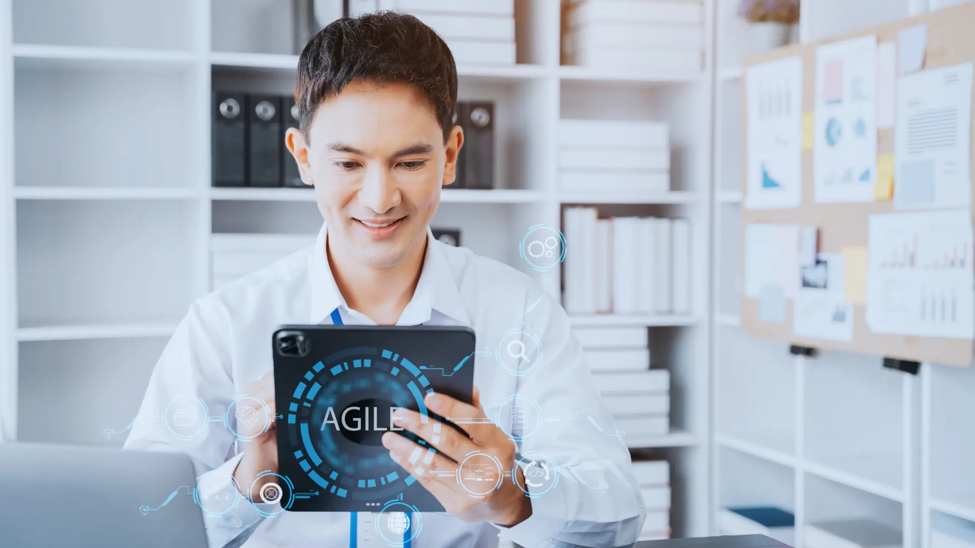 A man in a white shirt smiles while using a tablet at a desk. The word "AGILE" and digital interface graphics are superimposed, hinting at technology or what is an agile environment in software development. Shelves with books and files fill the background.